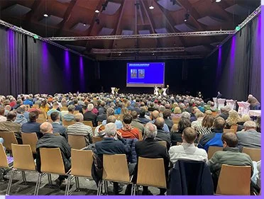 Menschen sitzten in einem Veranstaltungsraums und blicken auf eine große Leinwand. Die Atmosphäre ist ruhig und konzentriert.
